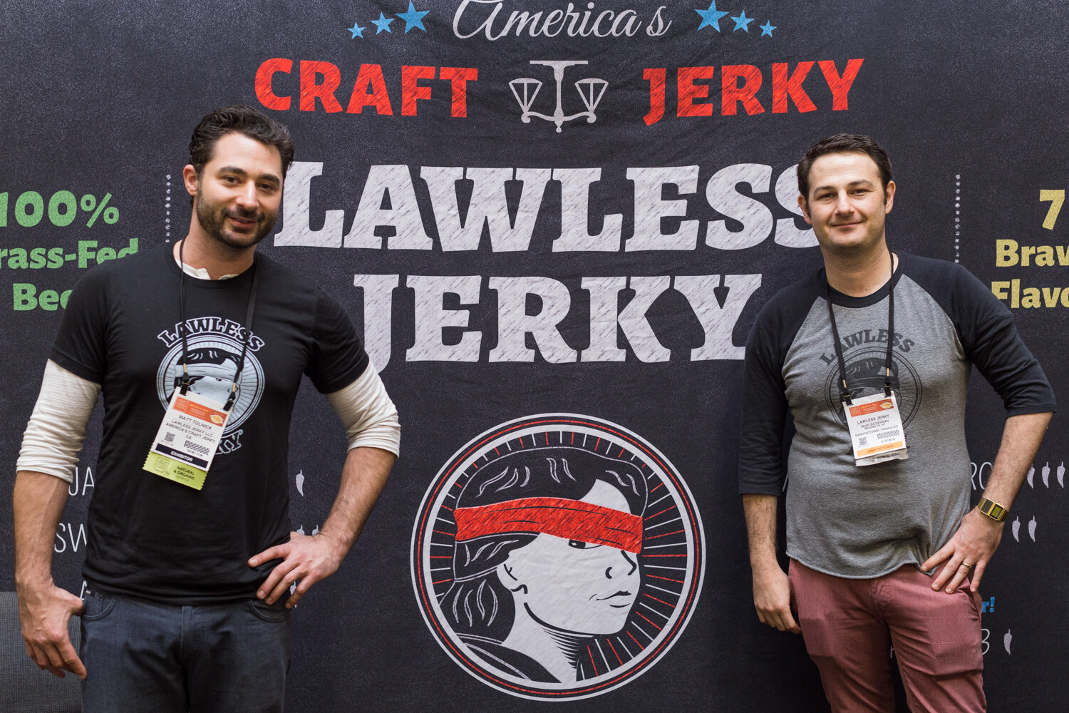 Two men with lanyards stand in front of a black backdrop reading America's Craft Jerky, Lawless Jerky with a blindfolded figure logo.