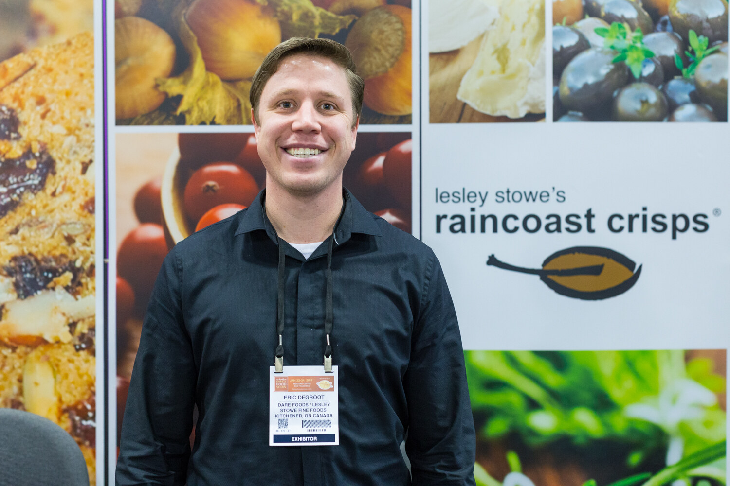 A man in a dark shirt with a lanyard stands in front of food photography panels and a sign reading lesley stowe's raincoast crisps.