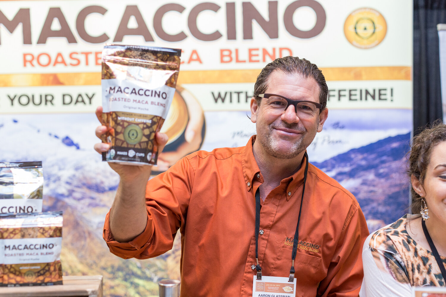A smiling man in an orange shirt holds up a bag of Macaccino Roasted Maca Blend in front of a Macaccino banner with mountain scenery.