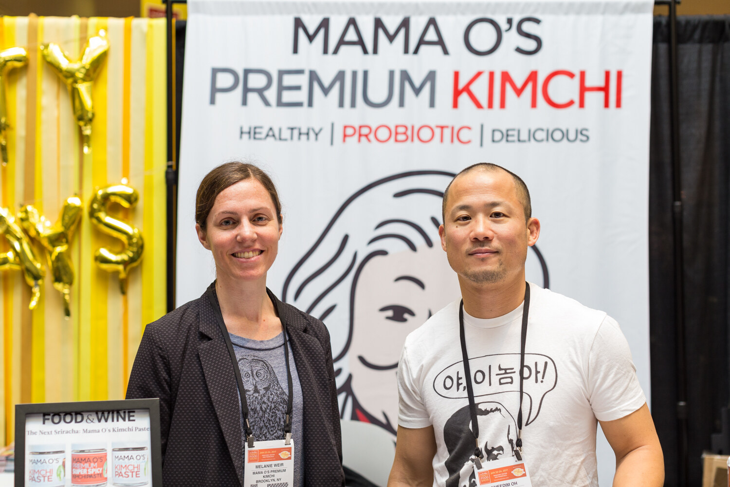 A woman in a black blazer and a man in a white t-shirt stand in front of a banner reading Mama O's Premium Kimchi, Healthy, Probiotic, Delicious.