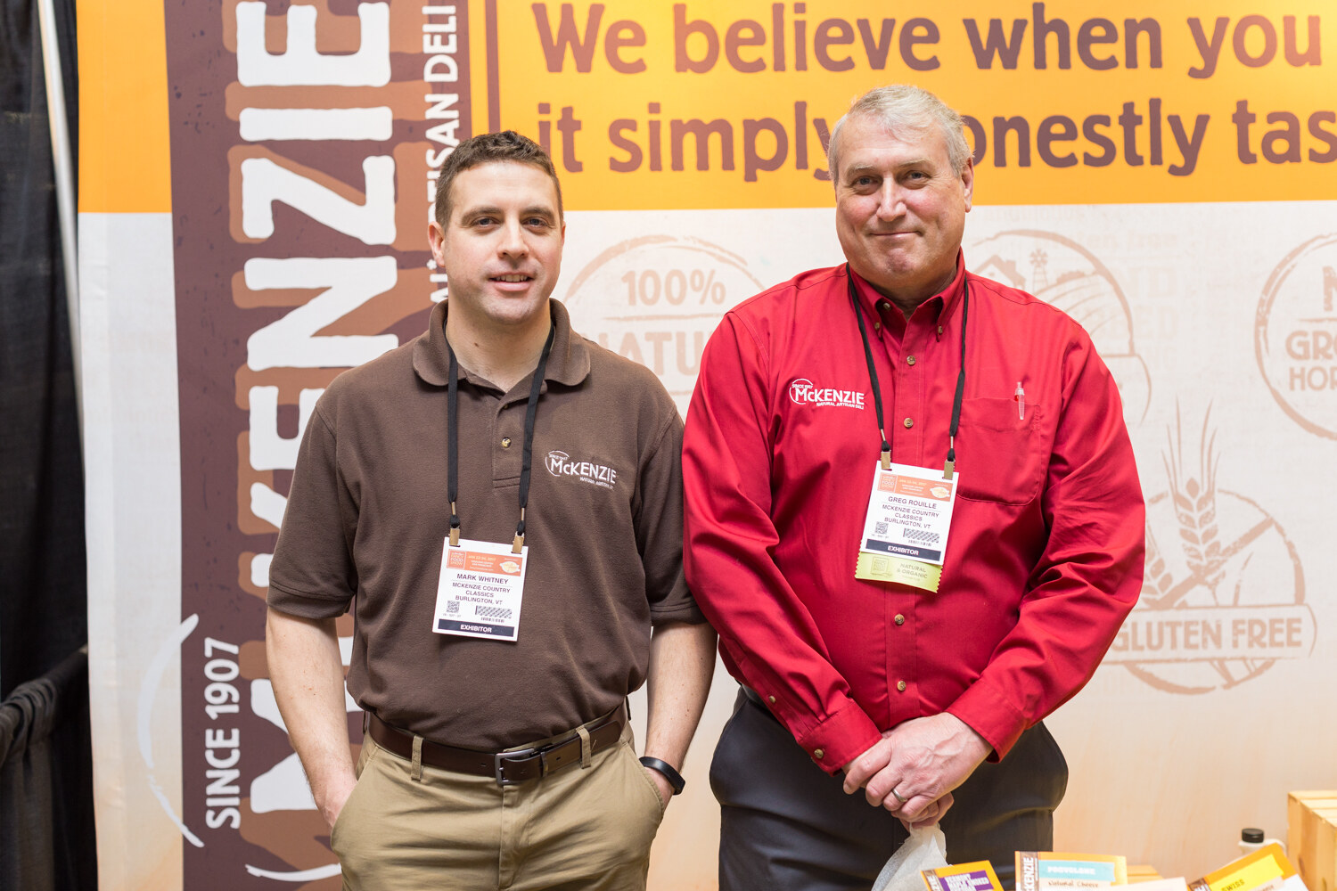 Two men in brown and red McKenzie shirts stand in front of a banner with McKenzie, Since 1907 and Gluten Free graphics.
