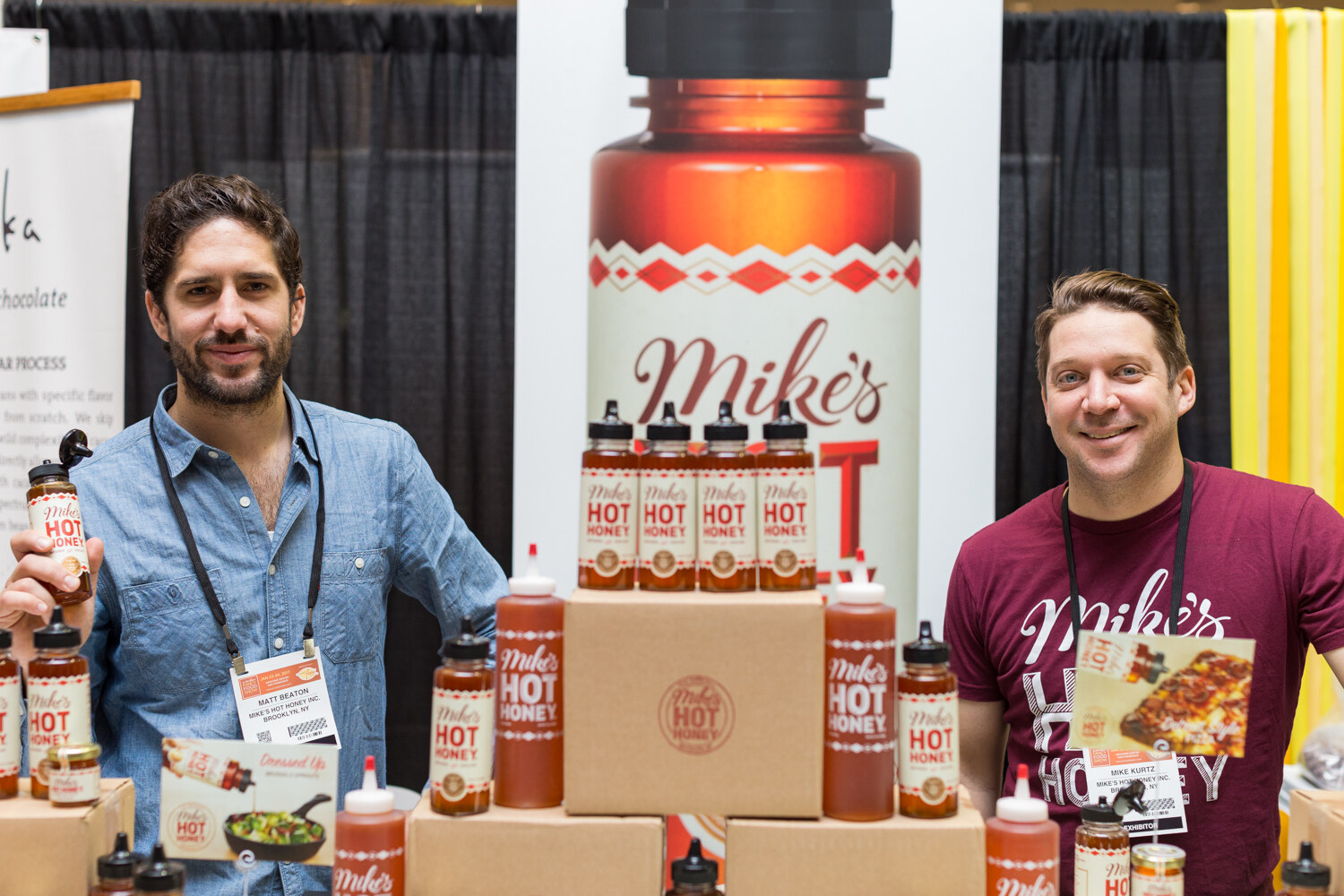 Two men stand behind boxes stacked with bottles of Mike's Hot Honey, one holding a squeeze bottle, in front of a large Mike's banner.
