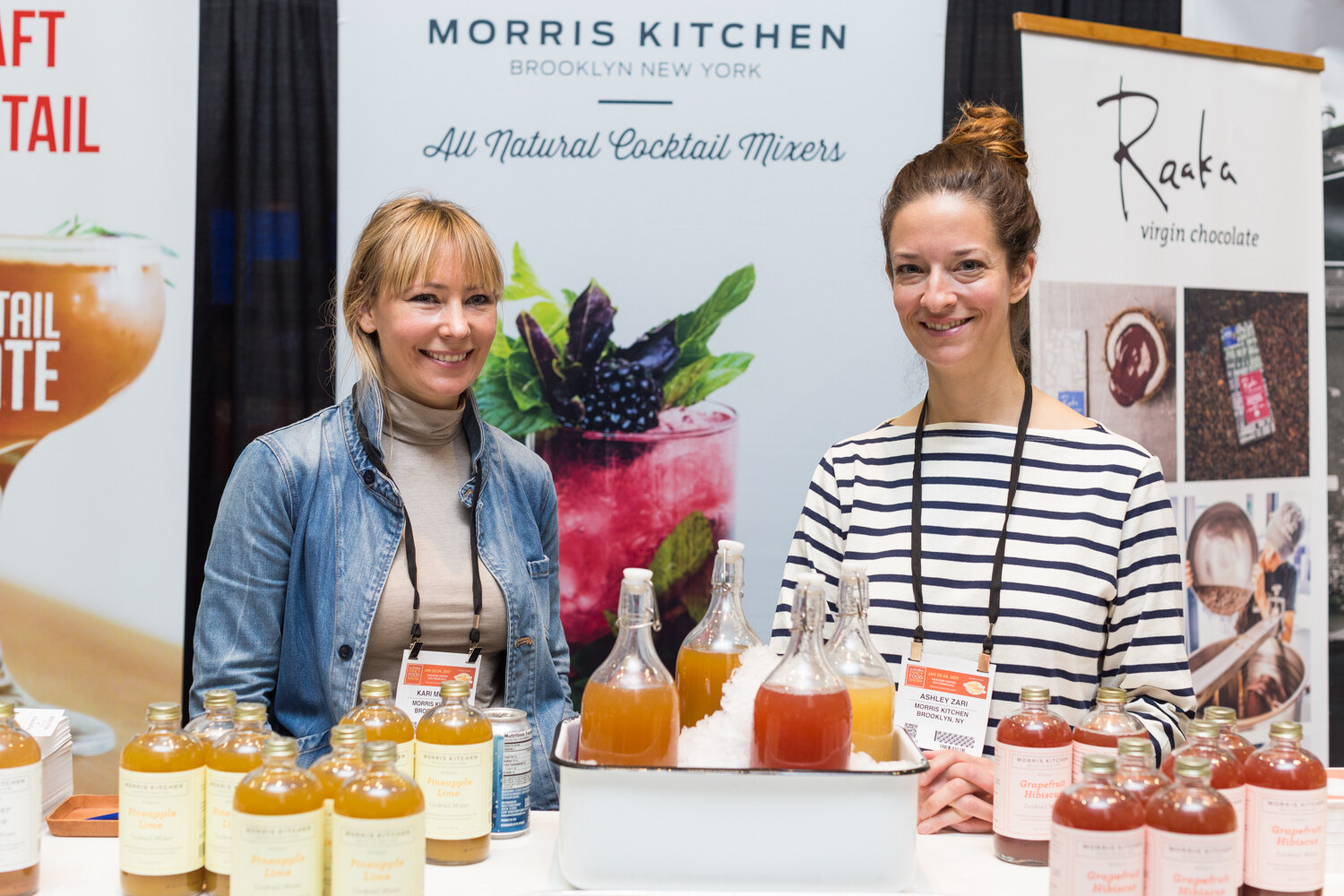 Two women with lanyards stand behind bottles of Pineapple Lime and Grapefruit Hibiscus mixers under a Morris Kitchen Brooklyn banner.