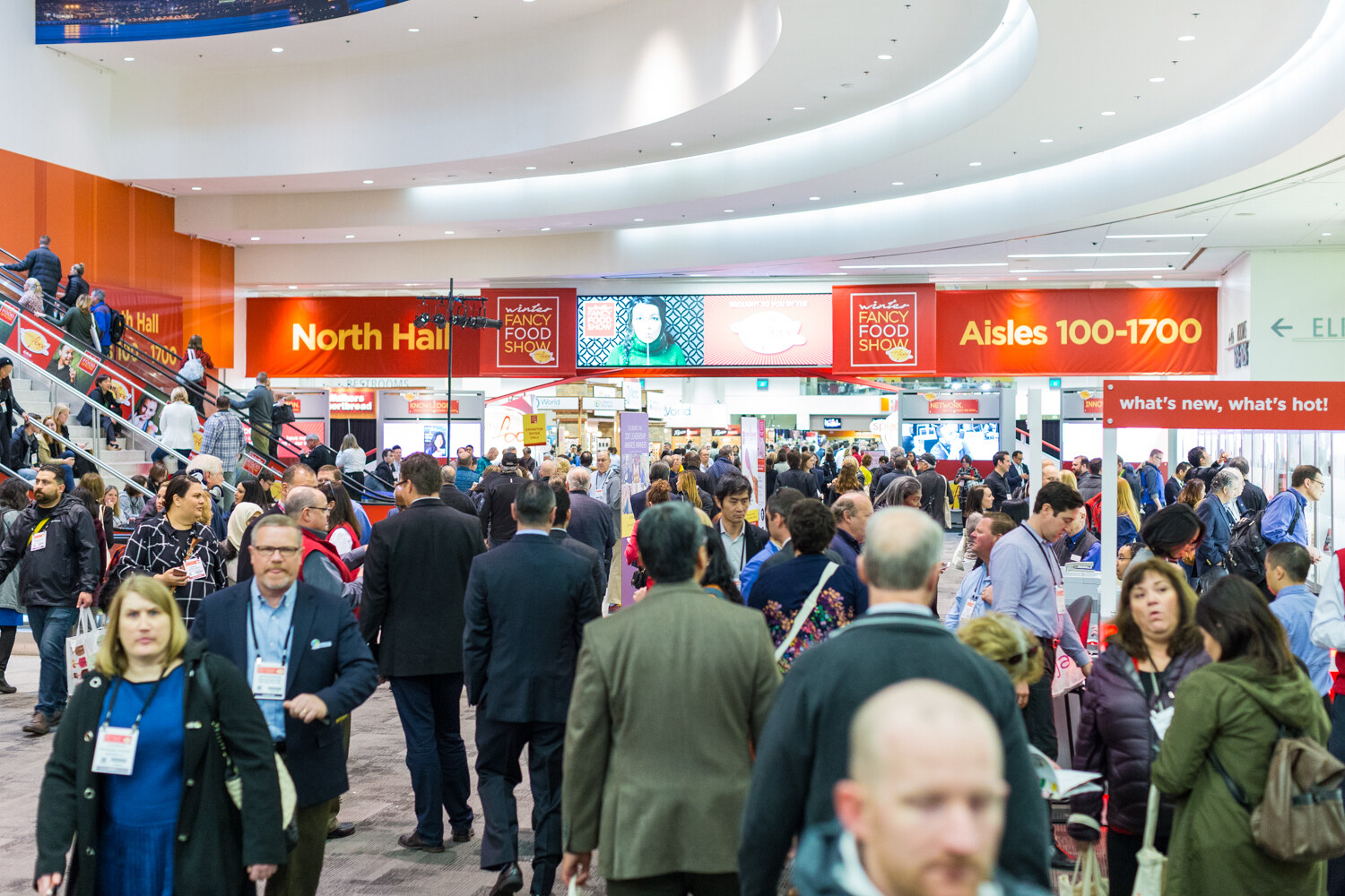 A busy concourse with orange signs reading North Hall and Aisles 100-1700, escalators on the left and many attendees walking toward the camera.