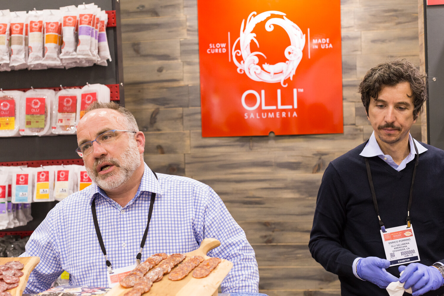 A man in a checked shirt speaks while a second man in blue gloves looks down, with a board of sliced salami in front and an orange Olli Salumeria sign behind.