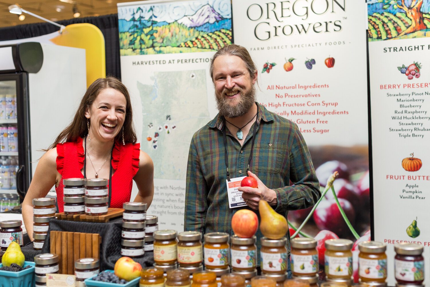 A woman in a red top and a bearded man holding an apple stand behind jars of preserves and fresh pears and apples in front of Oregon Growers banners.