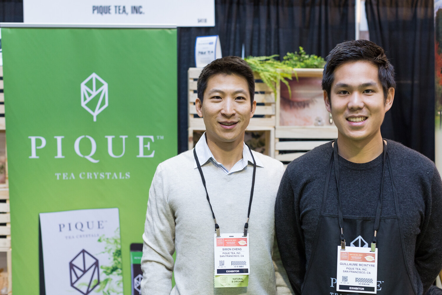 Two men with exhibitor badges stand in front of a green Pique Tea Crystals banner and wooden crates with plants.