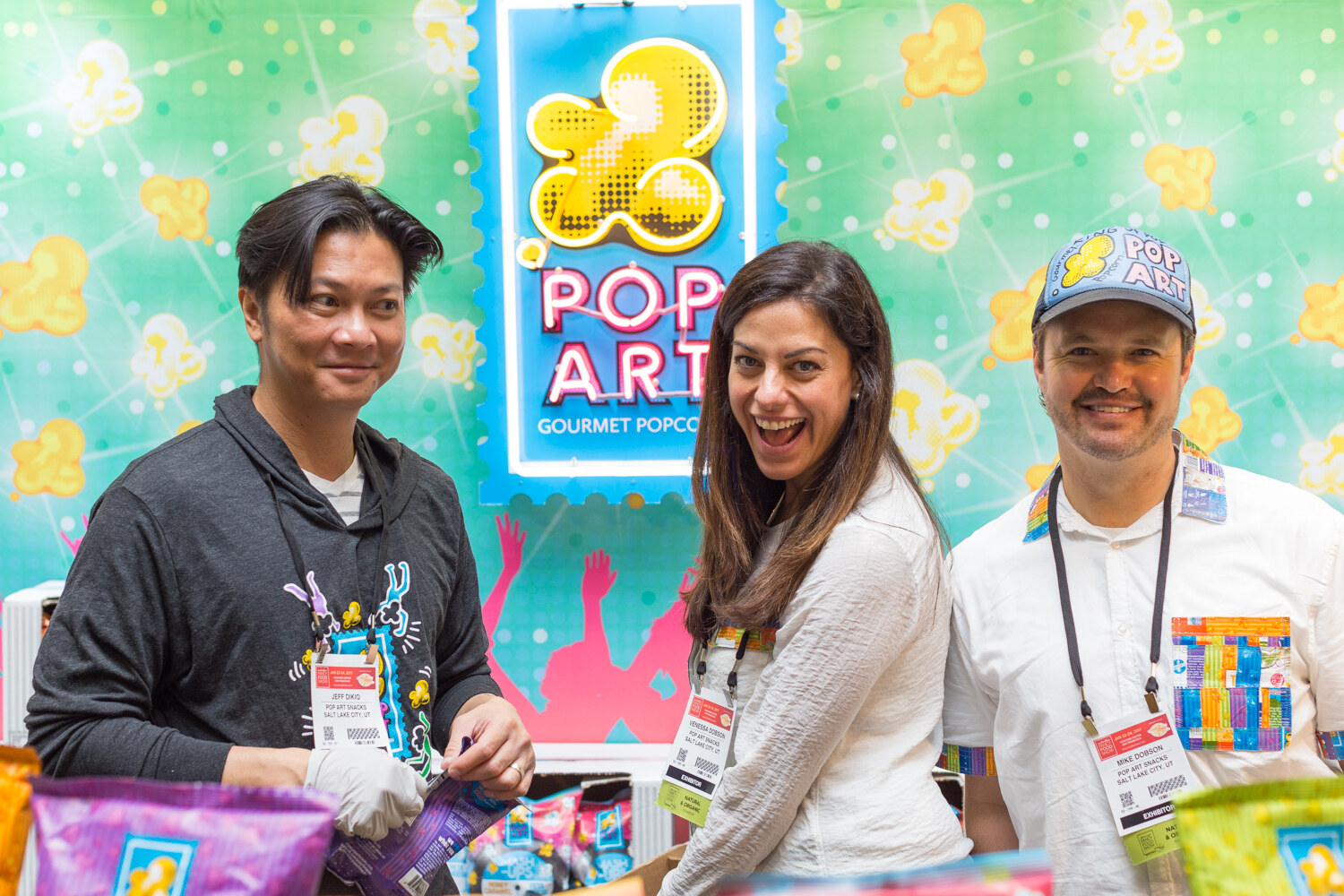 Two men and a woman stand in front of a green popcorn-patterned wall and a neon sign reading Pop Art Gourmet Popcorn, with bags of popcorn in the foreground.