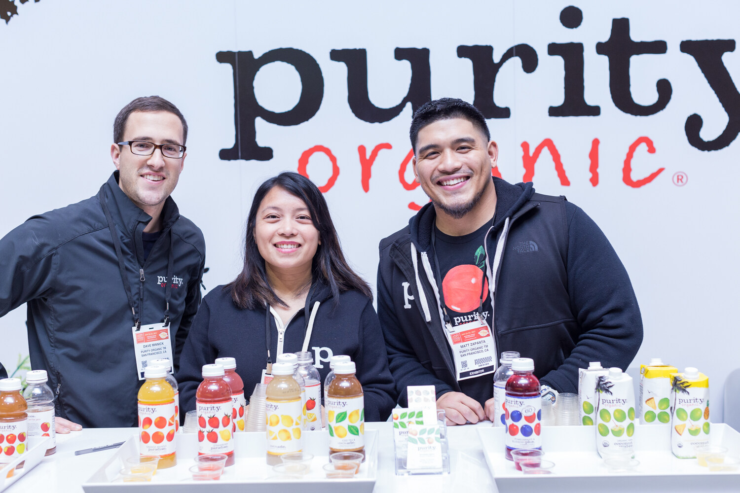 Three people in dark jackets stand behind a white counter of Purity Organic juice bottles and sample cups under a large Purity Organic sign.