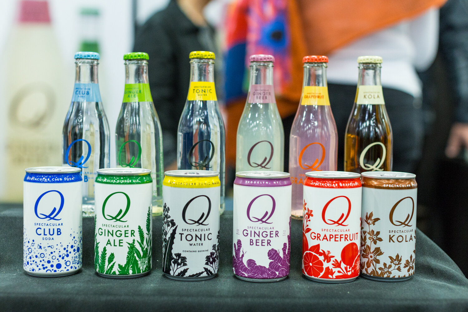 Six glass bottles and six small cans of Q Club Soda, Ginger Ale, Tonic Water, Ginger Beer, Grapefruit and Kola lined up on a table.