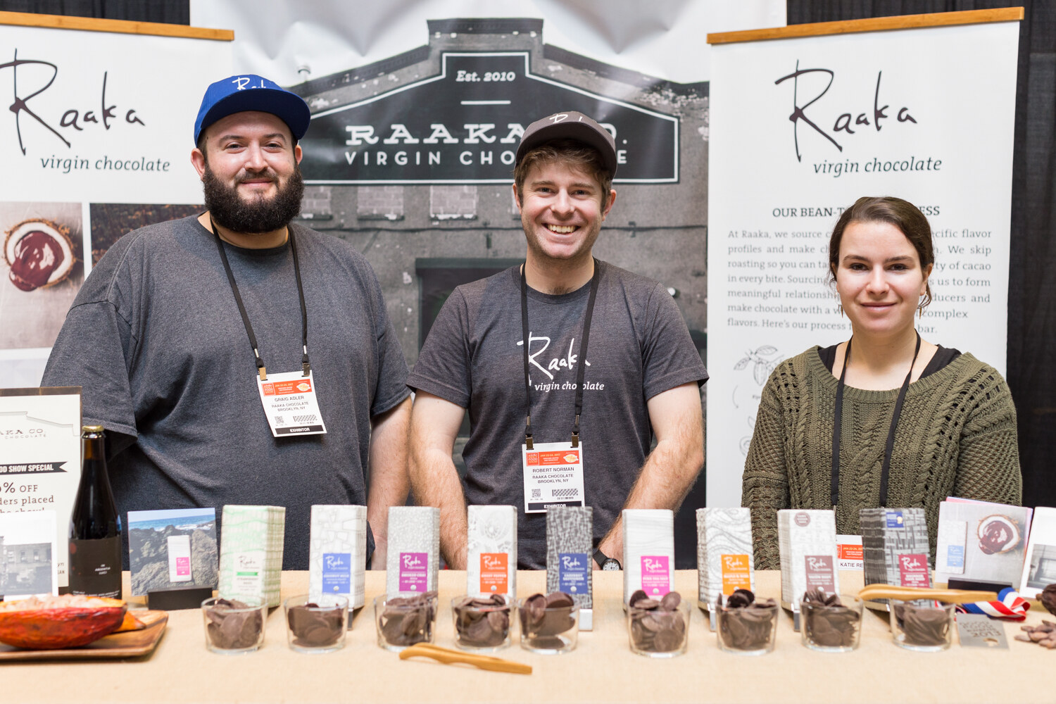 Two men and a woman stand behind a counter lined with Raaka chocolate bars and cups of chocolate pieces under Raaka virgin chocolate banners.