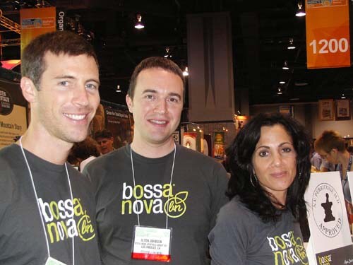 Two men and a woman in gray Bossa Nova T-shirts pose together at a booth, smiling at the camera, with an orange sign reading 1200 behind them.