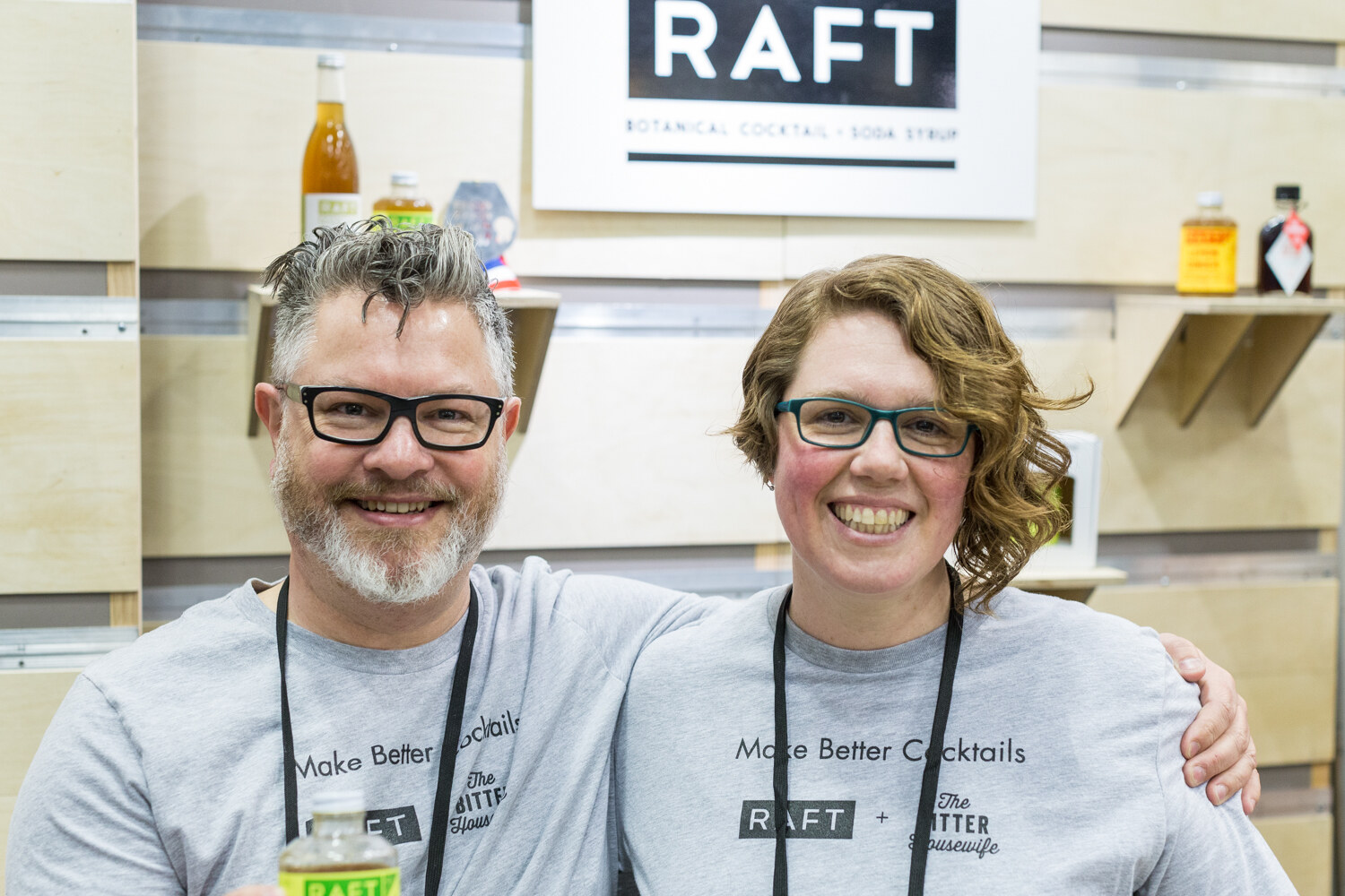 A man and a woman in gray Raft and The Bitter Housewife shirts smile under a Raft Botanical Cocktail and Soda Syrup sign.