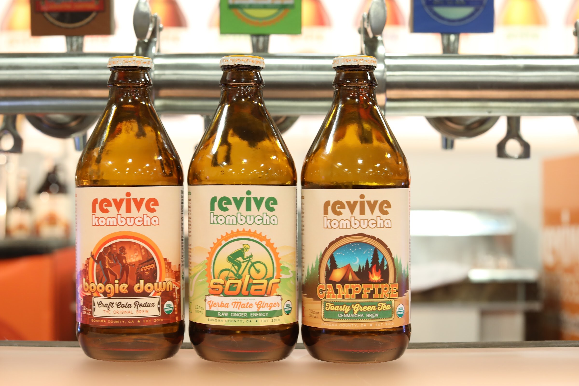 Three amber bottles labeled Revive Kombucha in Boogie Down, Solar and Campfire flavors in front of a row of tap handles.