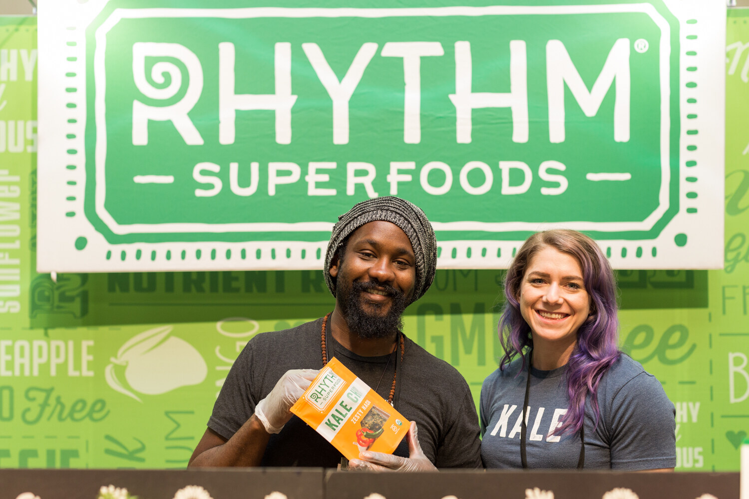 A man in a knit cap holding a bag of Rhythm kale chips and a woman with purple hair smile beneath a green Rhythm Superfoods sign.