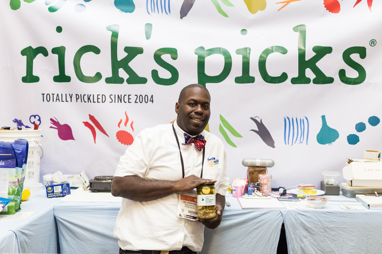 A smiling man in a white chef coat and red bow tie holds a jar of pickles in front of a banner reading rick's picks, Totally Pickled Since 2004.
