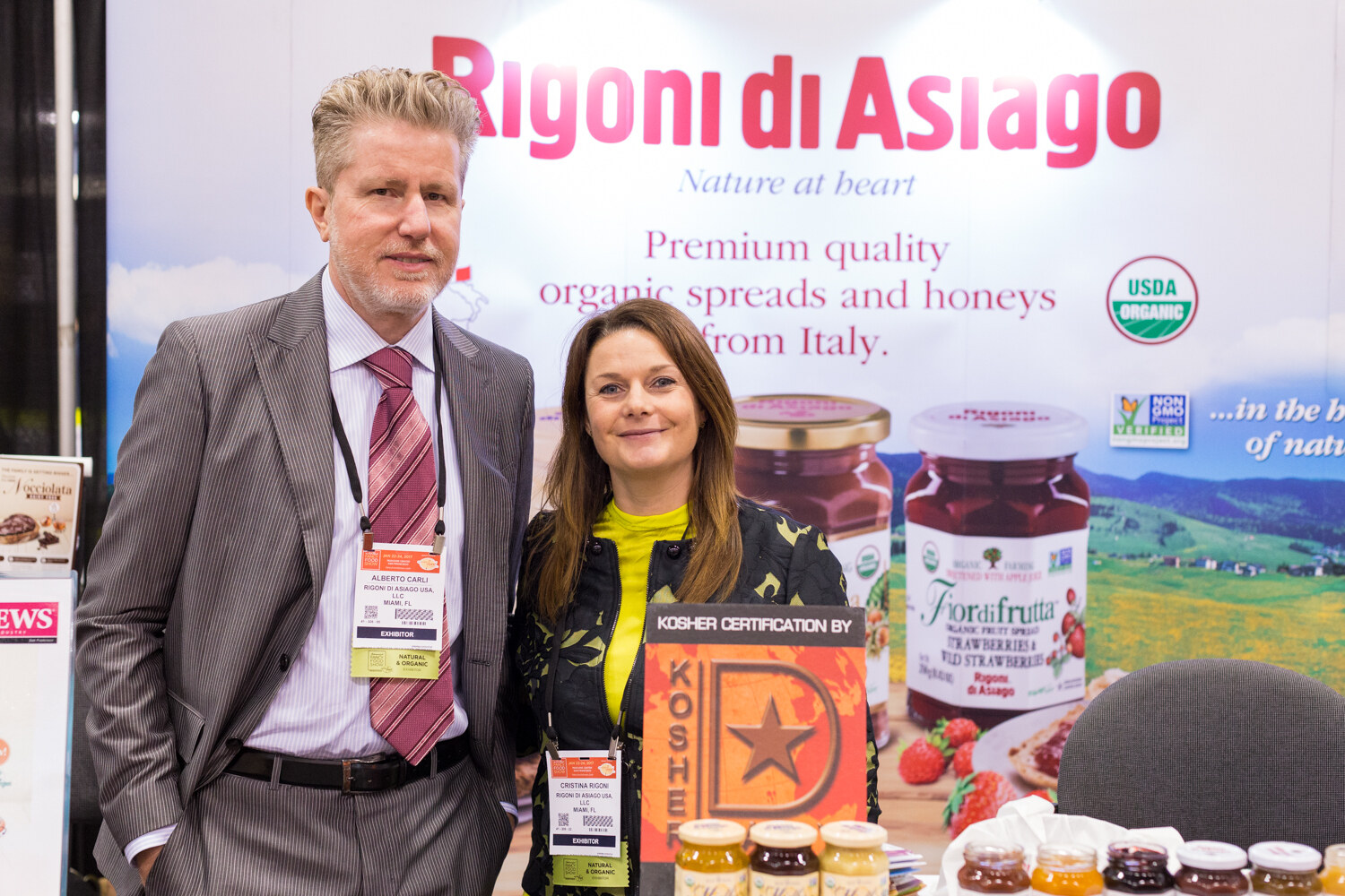 A man in a gray suit and a woman in a green jacket stand in front of a banner reading Rigoni di Asiago, Premium quality organic spreads and honeys from Italy.