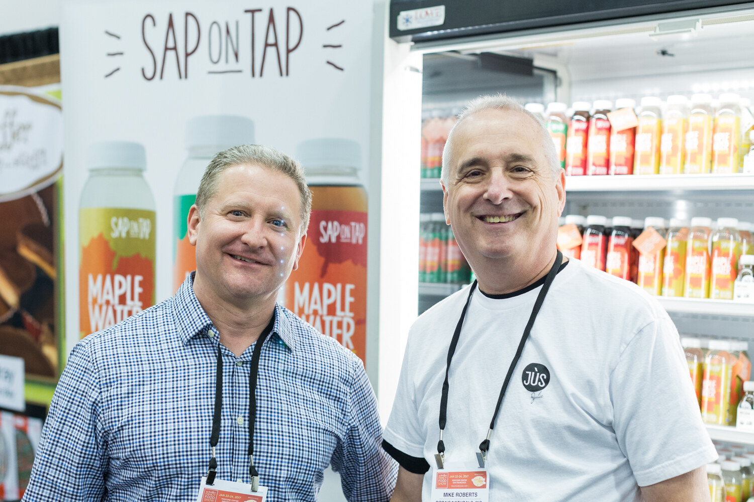 Two men with lanyards stand in front of a Sap on Tap Maple Water banner and a refrigerated case of bottled juices.