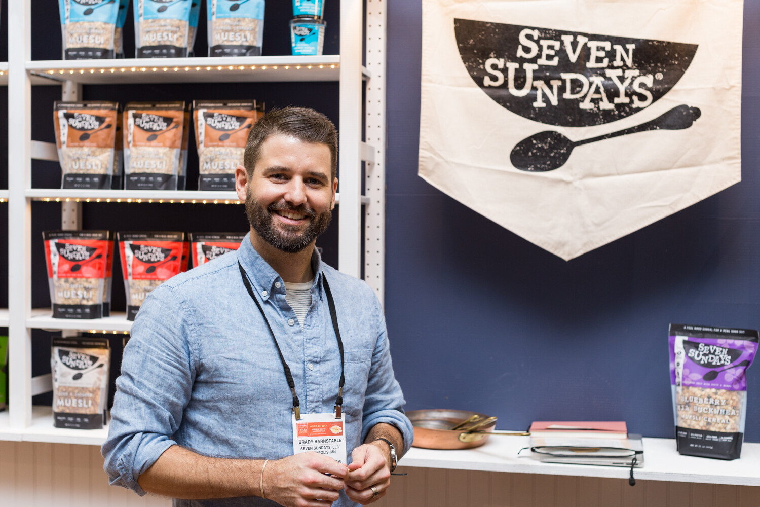 A bearded man in a blue shirt smiles in front of shelves of Seven Sundays muesli bags and a cloth banner with the Seven Sundays bowl logo.