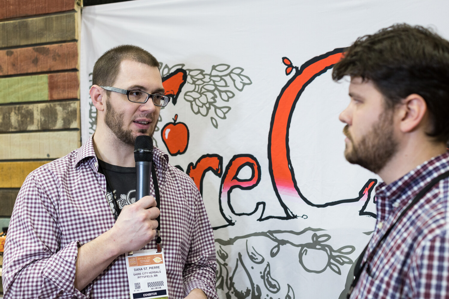 A bearded man with glasses speaks into a microphone while a second man in profile listens, in front of a white banner with red apple artwork.