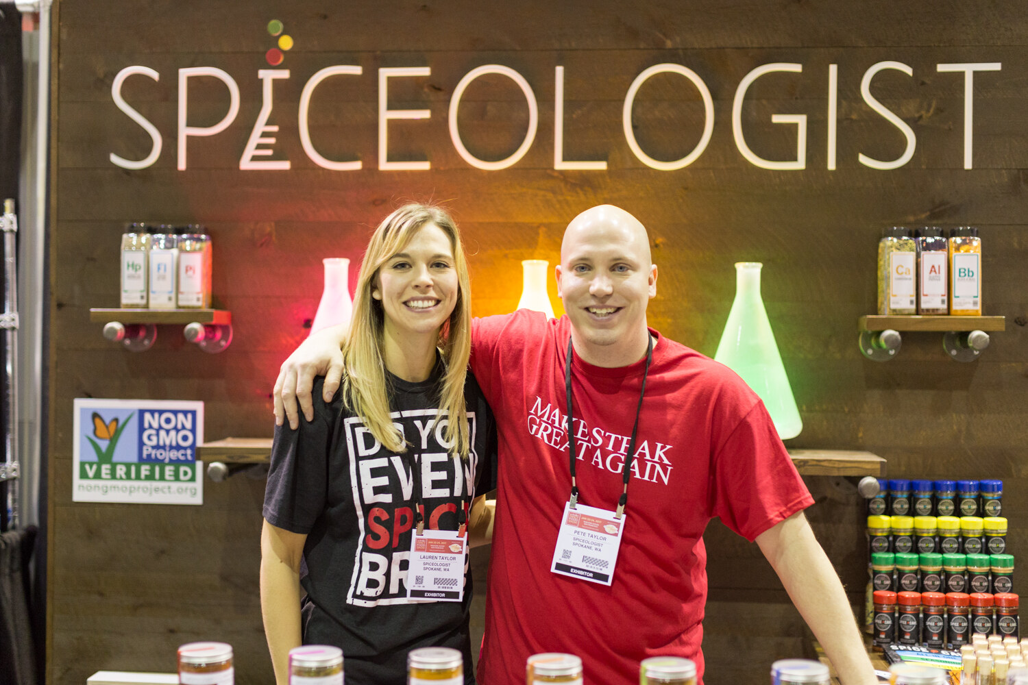 A woman and a man in a red Make Steak Great Again shirt stand in front of a wood wall with the Spiceologist sign, lit flasks and jars of spices.