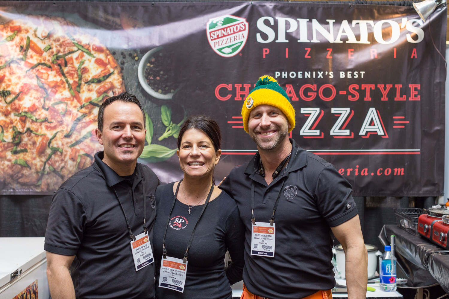 A man, a woman and a man in a yellow and green beanie stand in front of a banner reading Spinato's Pizzeria, Phoenix's Best Chicago-Style Pizza.
