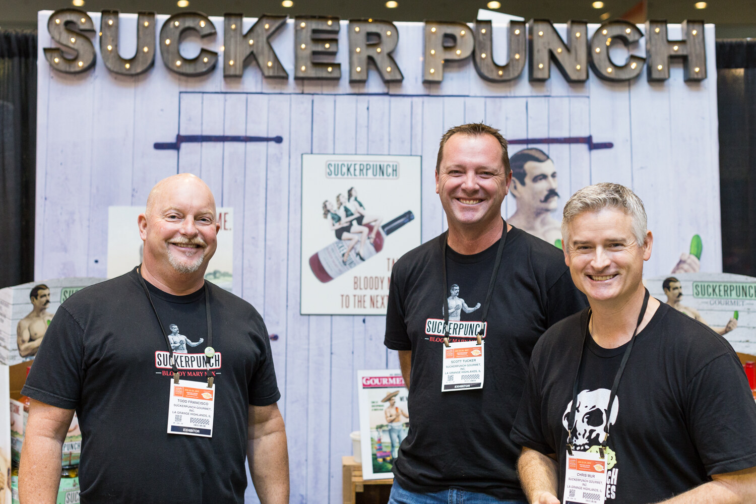 Three smiling men in black Suckerpunch shirts stand in front of a white plank wall under a lit marquee sign reading Sucker Punch.