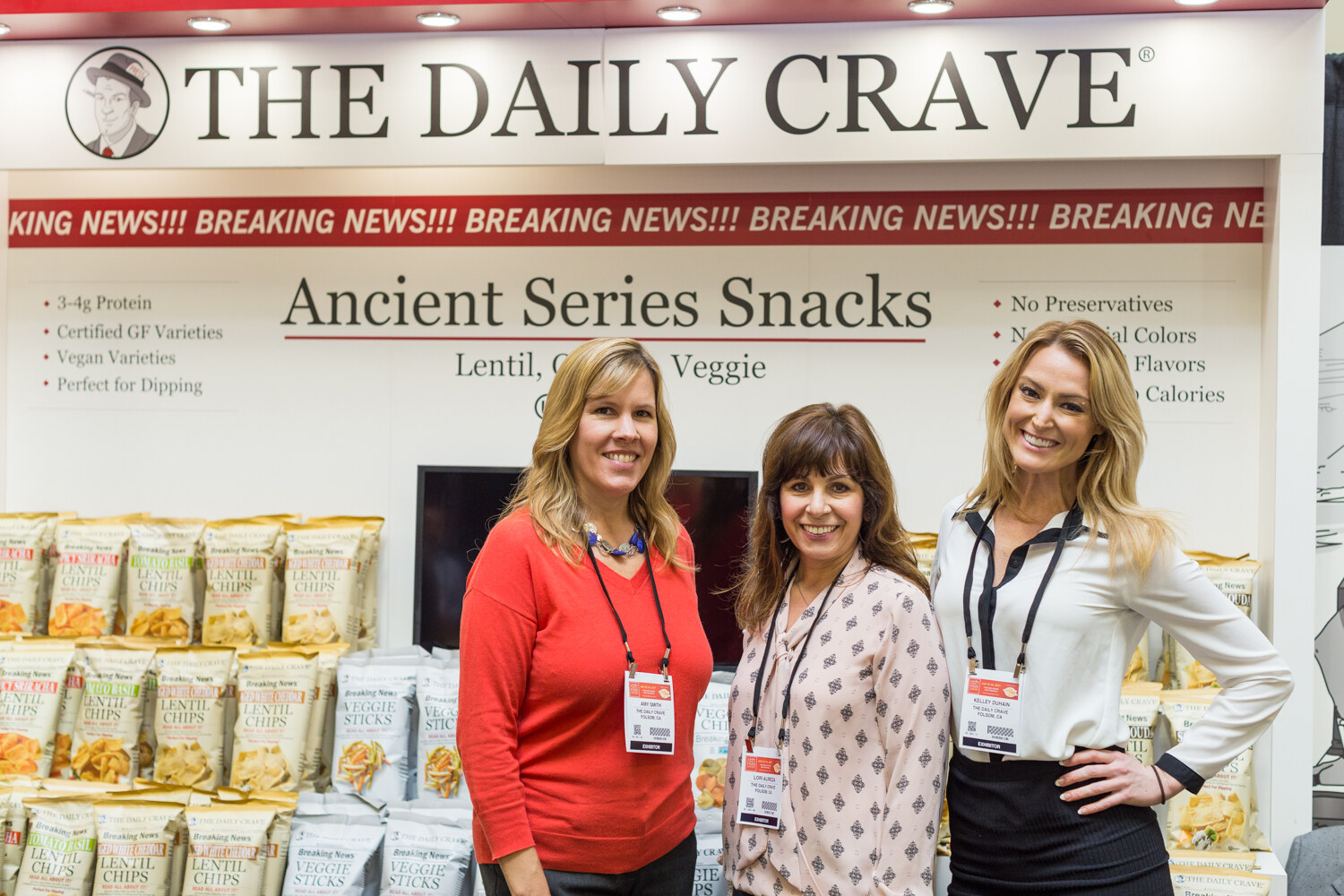 Three women with exhibitor badges stand in front of a The Daily Crave booth with a Breaking News banner and bags of lentil chips.