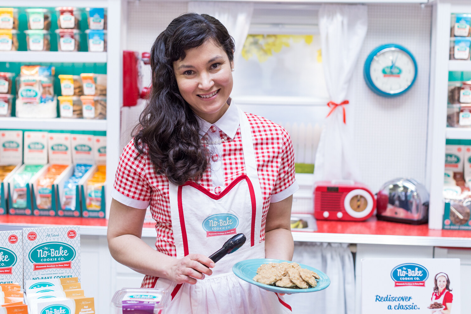 A smiling woman in a red gingham dress and white apron holds a plate of cookies and tongs in front of shelves of The No-Bake Cookie Co. packages.