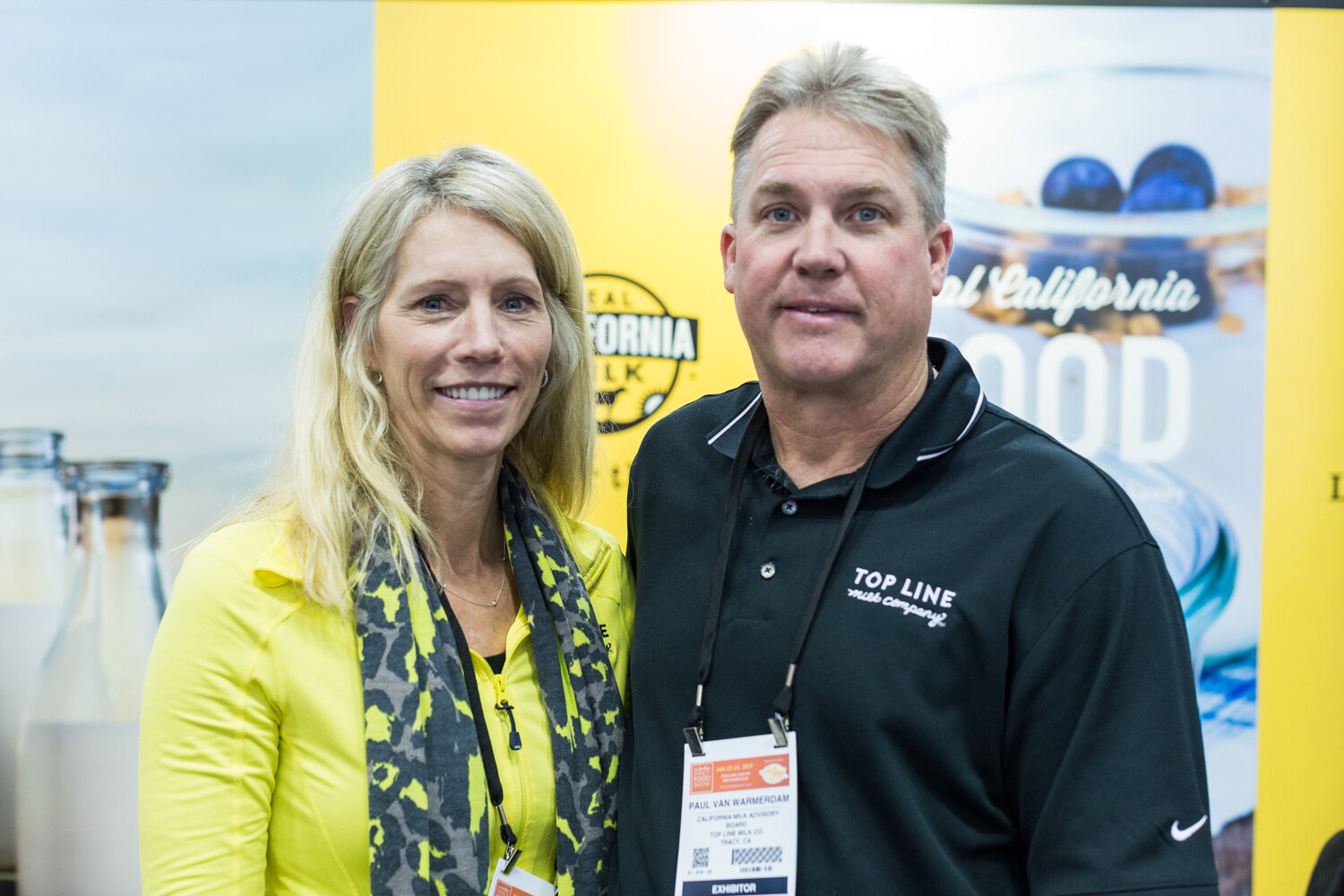 A woman in yellow and a man in a black Top Line Milk Company polo stand in front of a yellow banner with a Real California Milk logo and milk bottles.