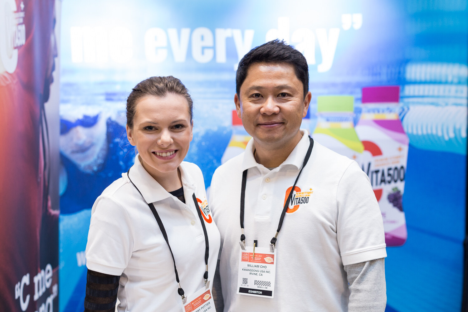Two smiling people in white polo shirts with Vita500 logos stand in front of a blue banner showing bottles of Vita500 drinks.
