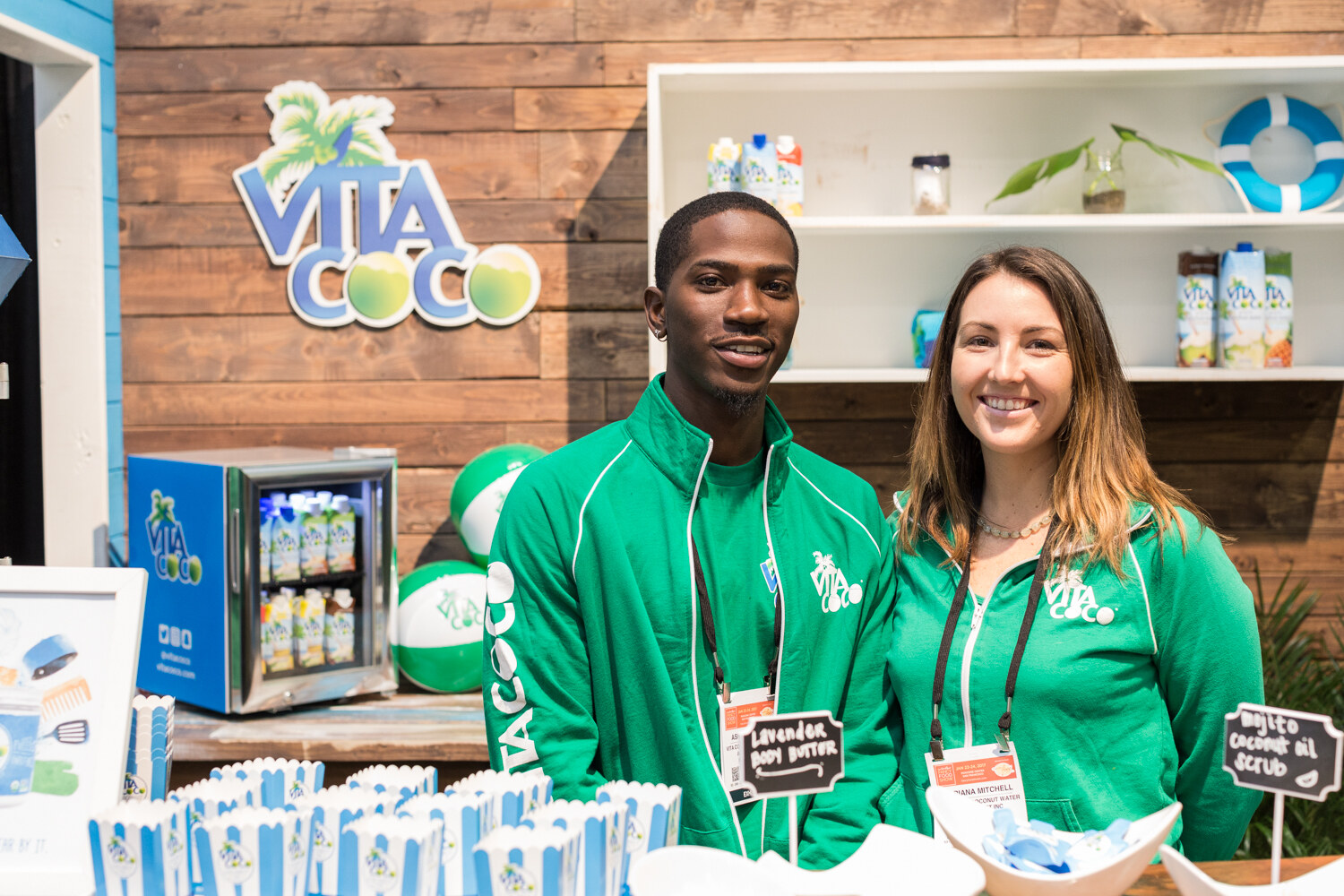 Two people in green Vita Coco jackets stand at a wooden booth with a Vita Coco logo sign, a small refrigerator of coconut water and beach balls.