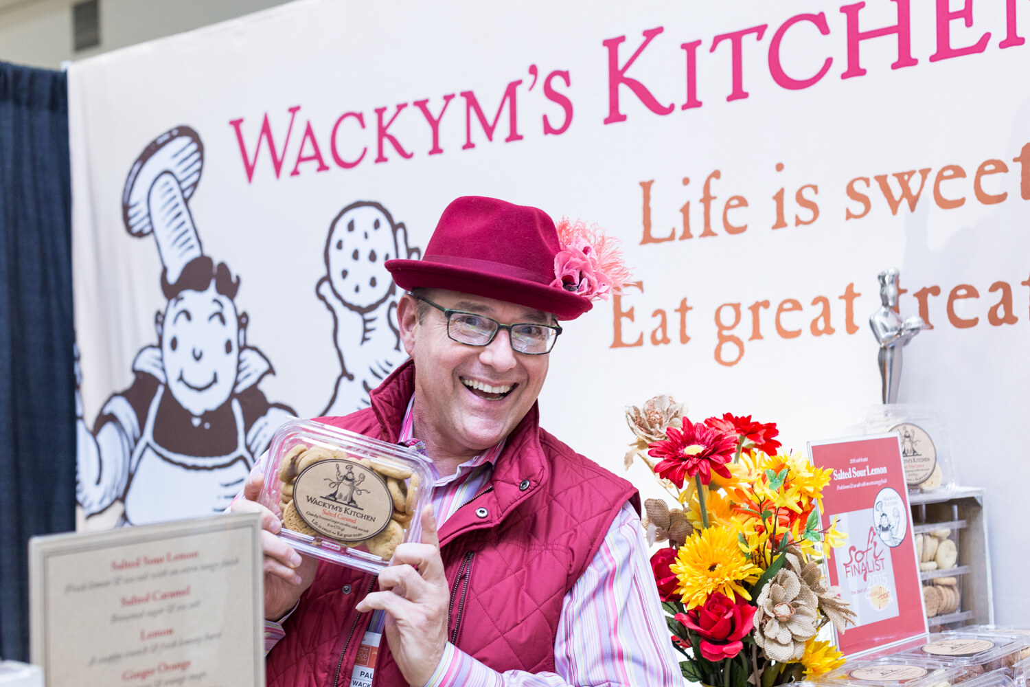 A laughing man in a burgundy hat and vest holds up a container of cookies in front of a Wackym's Kitchen banner with a cartoon baker.