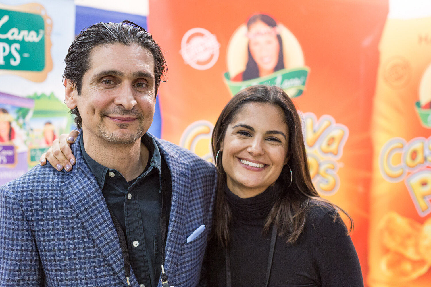 A man in a checked blazer and a woman in a black top stand arm in arm in front of orange Wai Lana cassava chips banners.