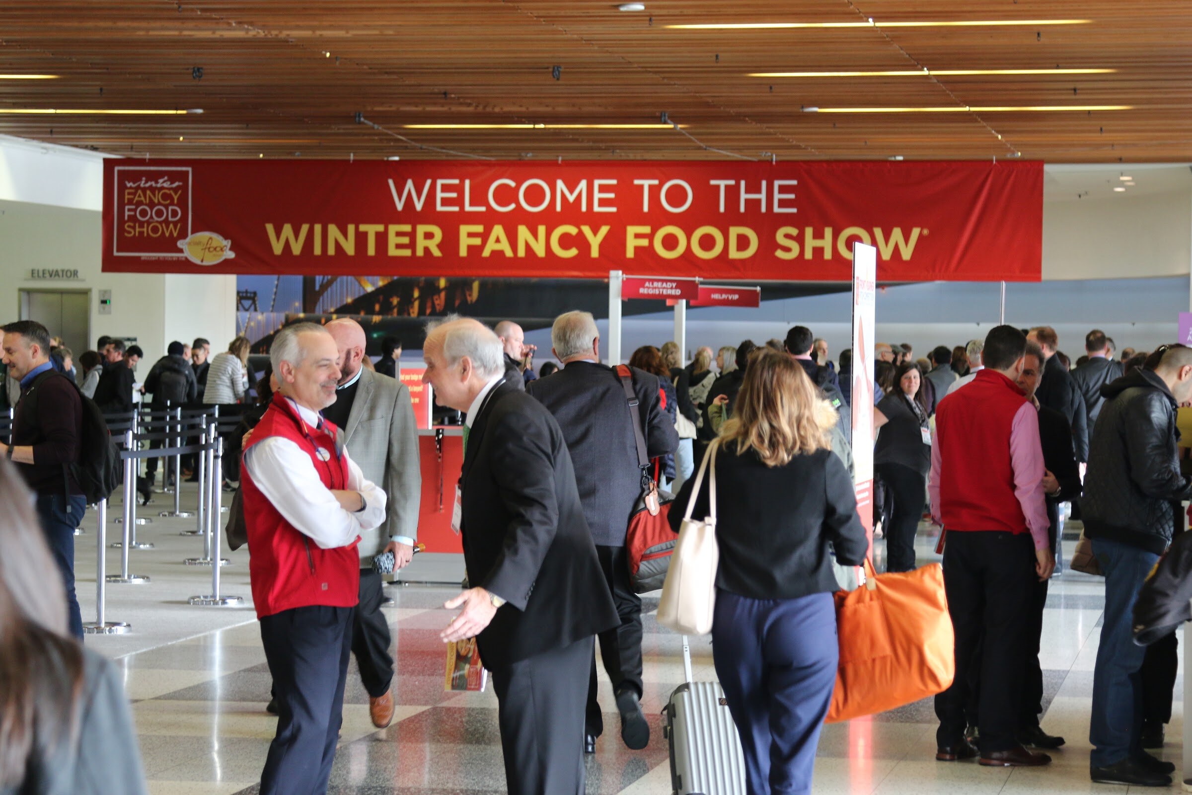 A red banner reading Welcome to the Winter Fancy Food Show hangs over a crowded entrance where people wait near registration signs, including a man in a red vest.