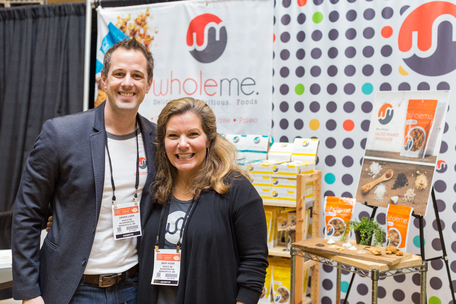 A man in a gray blazer and a smiling woman in a black cardigan stand in front of a Wholeme banner and a polka-dot wall with product displays.