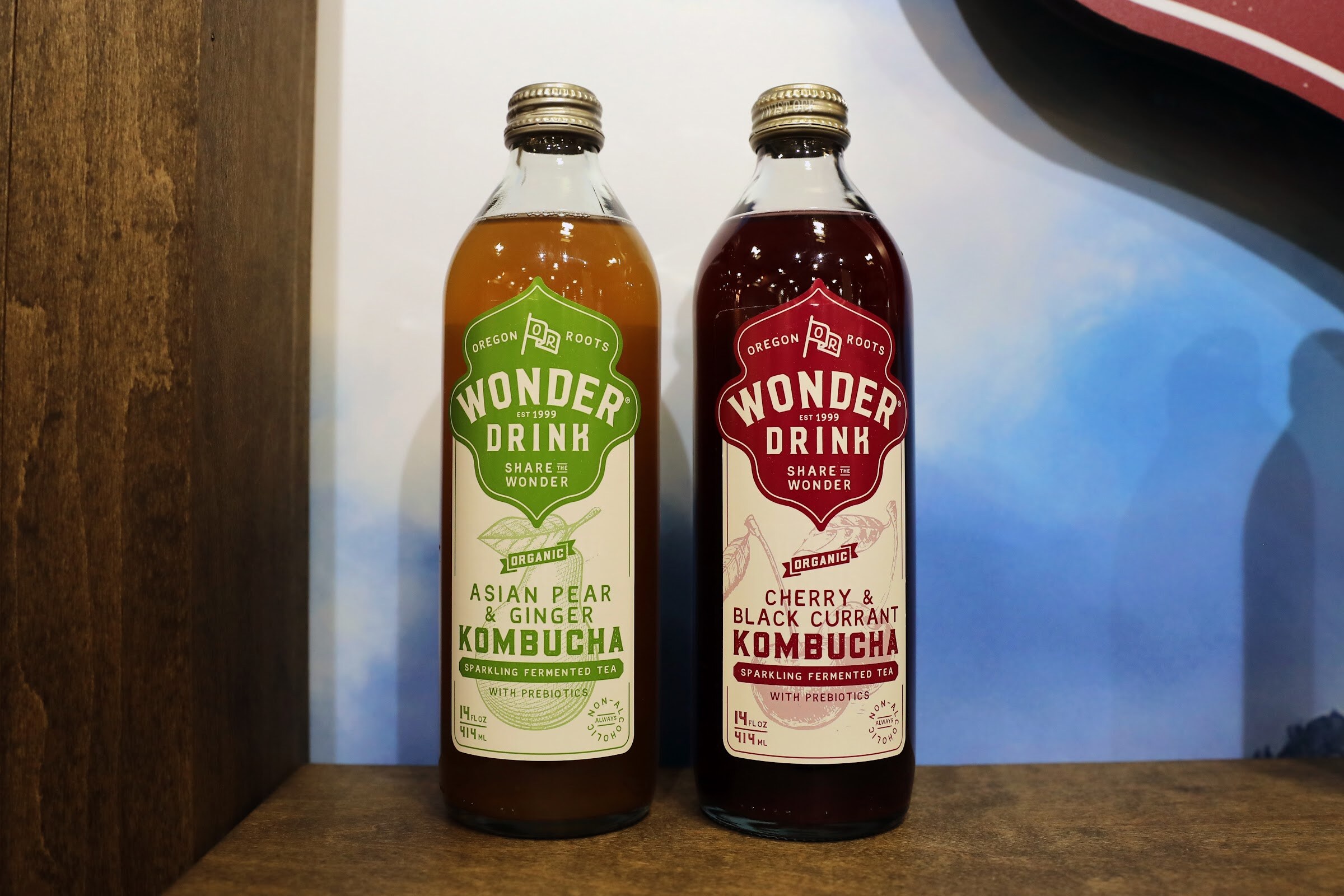 Two glass bottles of Wonder Drink kombucha, Asian Pear and Ginger and Cherry and Black Currant, on a wooden shelf.