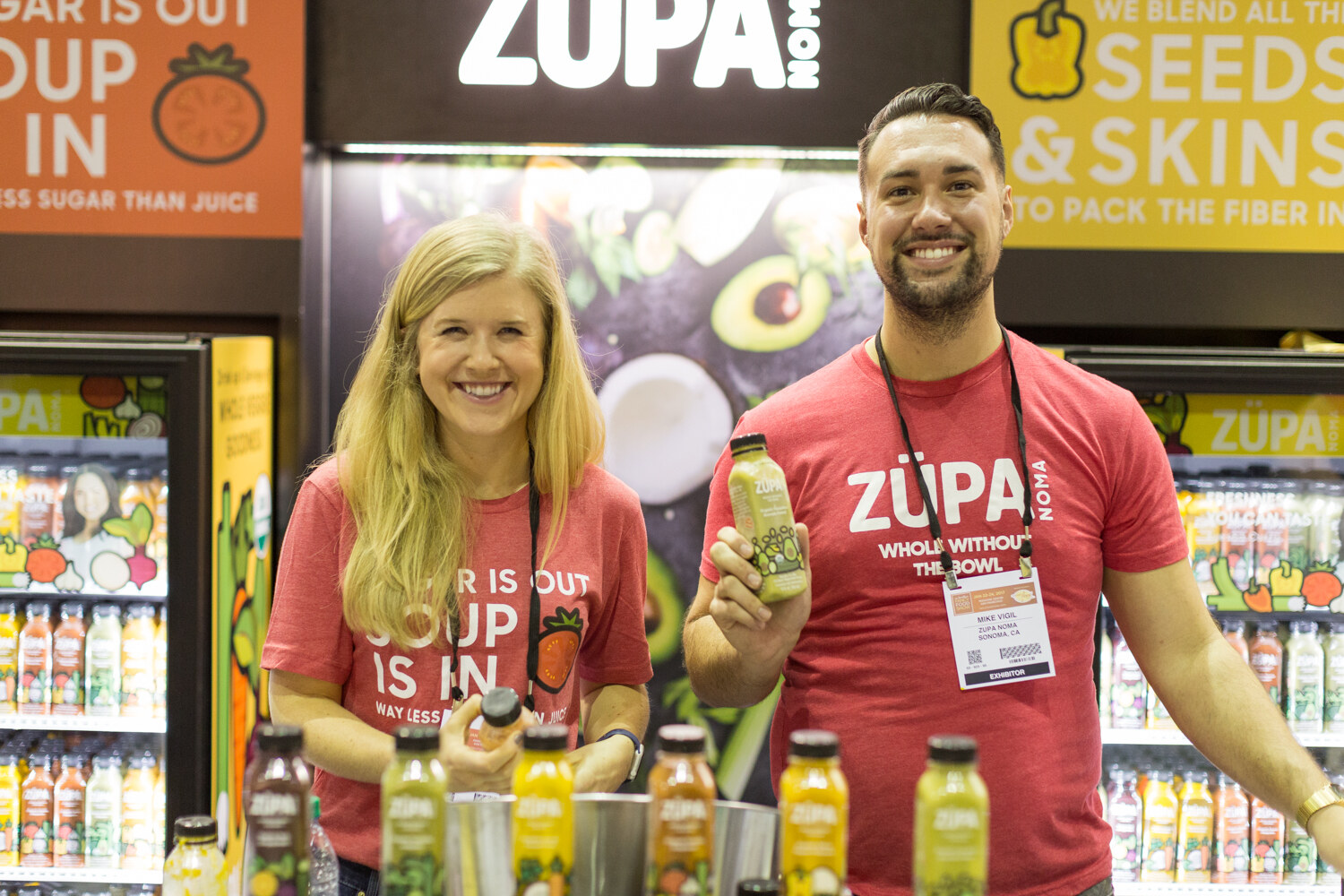 A woman and a man in red Zupa Noma shirts smile at a booth with refrigerated cases of bottled drinks, and the man holds up a green bottle.