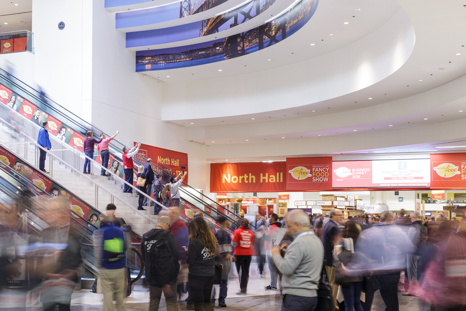 Crowd of attendees with badges on escalators and a walkway beneath North Hall and Fancy Food Show banners.