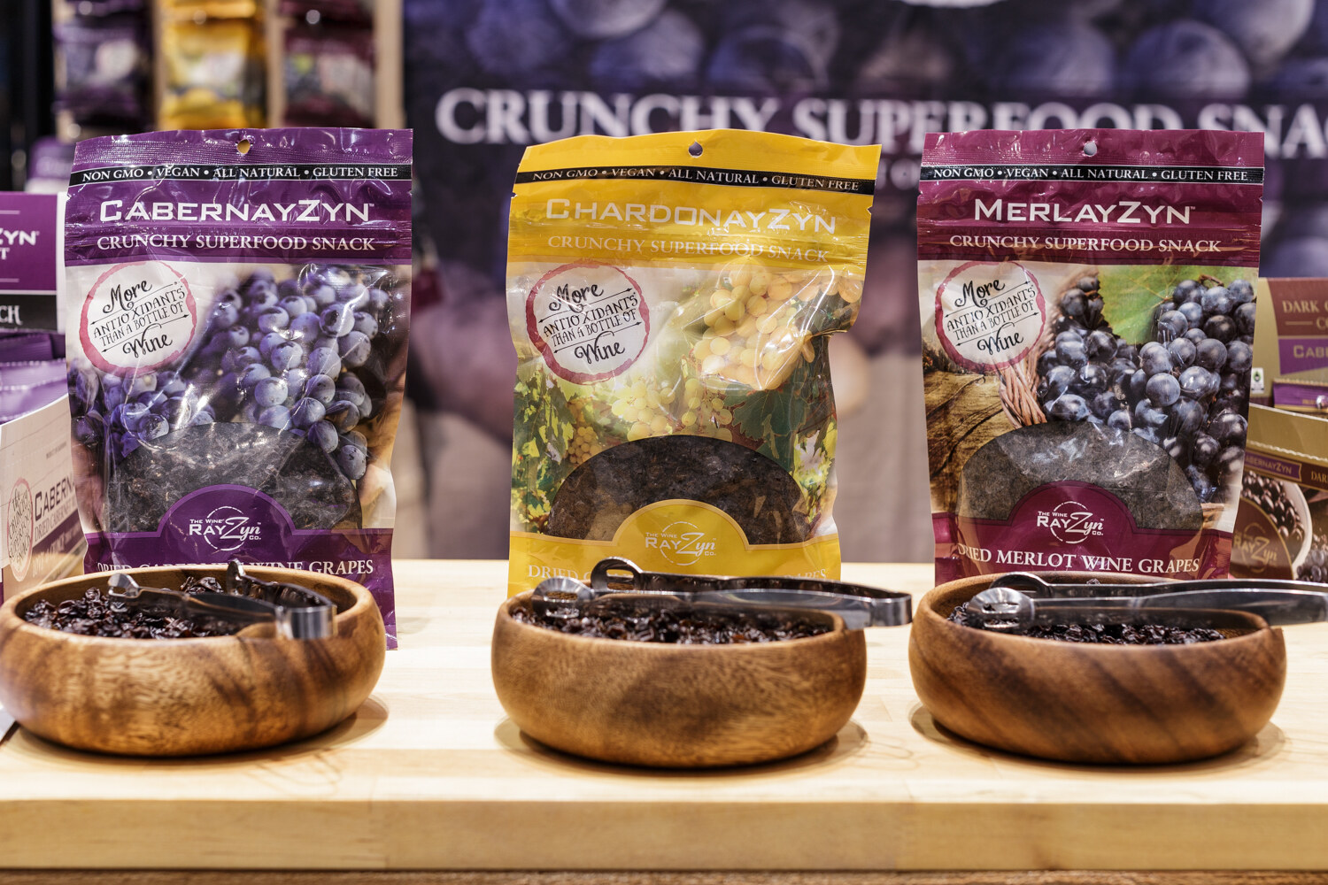 Bags of CabernayZyn, ChardonnayZyn and MerlayZyn above wooden bowls of dried grapes.