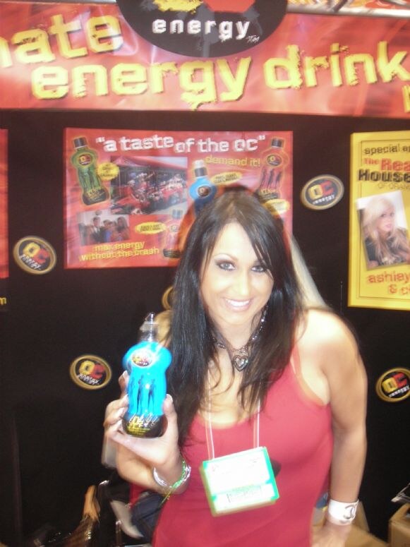 A smiling dark-haired woman in a red top and badge holds a blue OC Energy bottle in front of OC Energy posters and logos on a black booth wall.