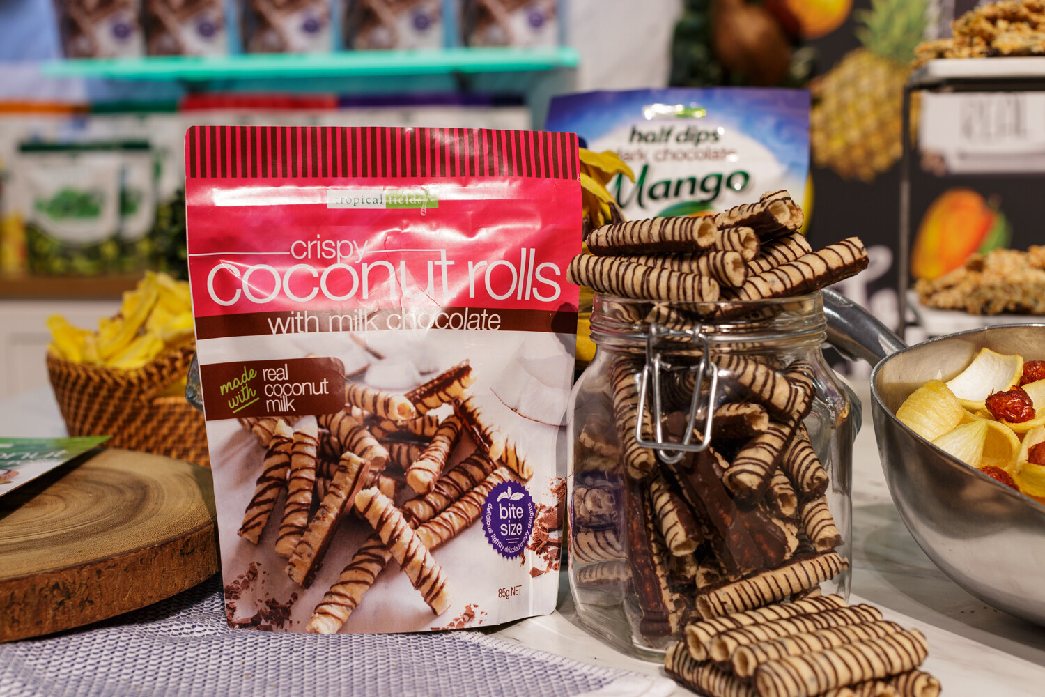 A pink bag of coconut rolls with a jar of striped wafer rolls and other snacks behind.