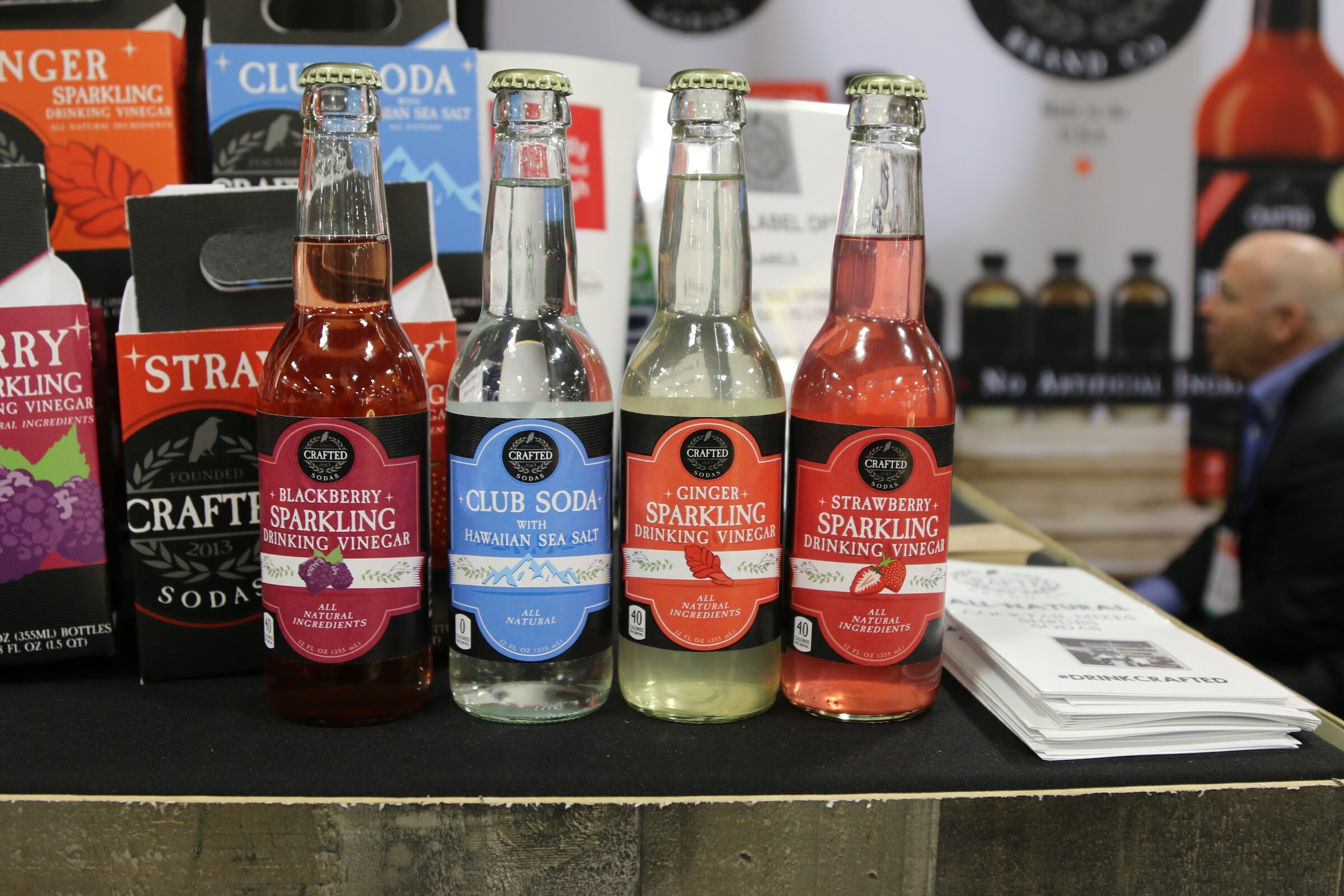 Four bottles of Crafted sparkling drinking vinegar and club soda in front of a sign.