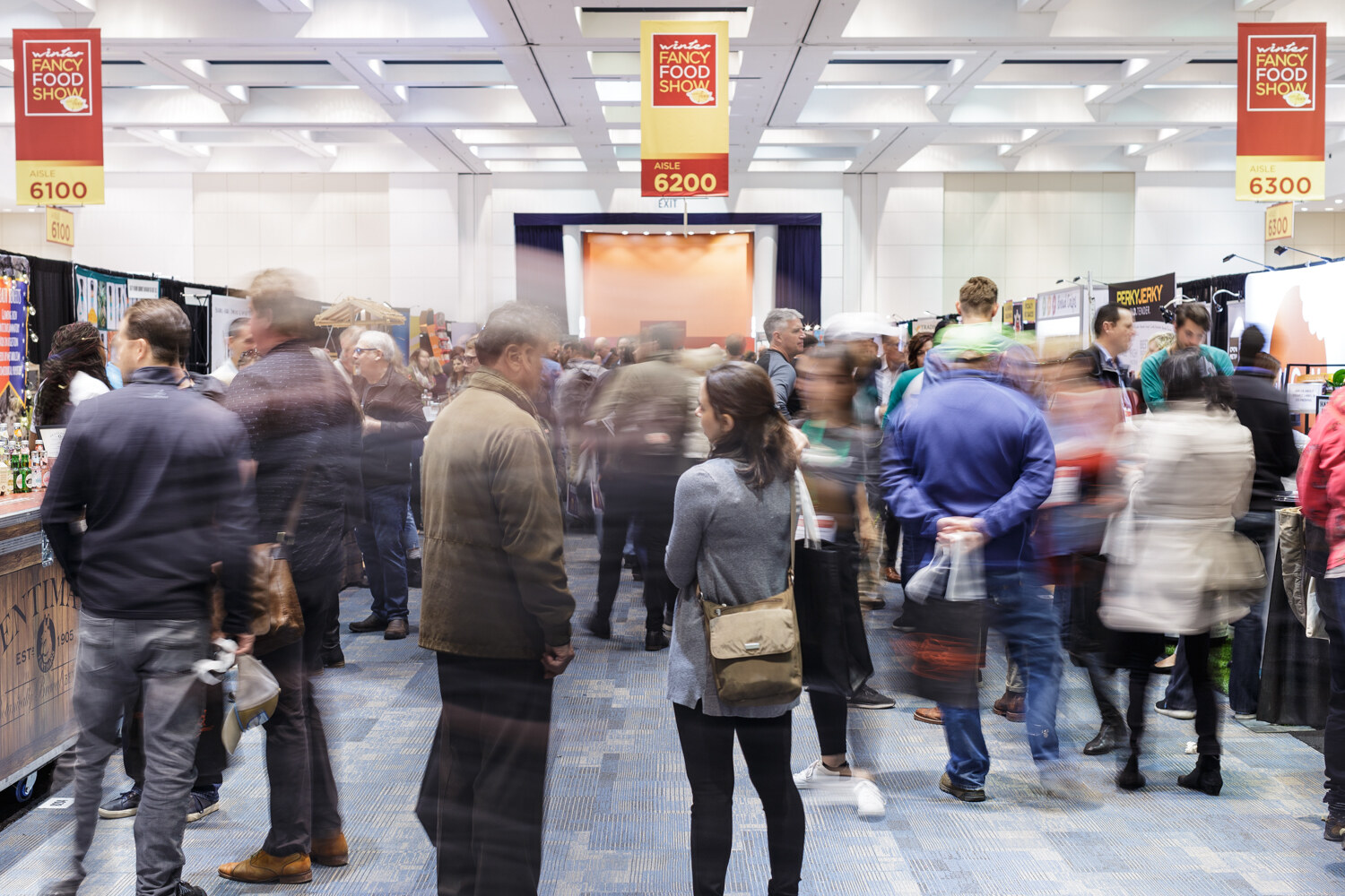 Wide view of a crowded exhibit-hall aisle with attendees in motion and booths on both sides.