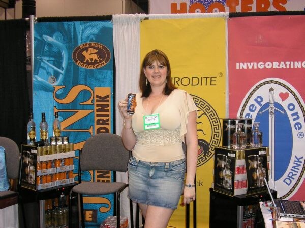 A woman in a cream top and denim skirt holds a can in front of Blue Jeans Energy Drink and Peep One banners, with bottles on display stands and a Hooters sign above.