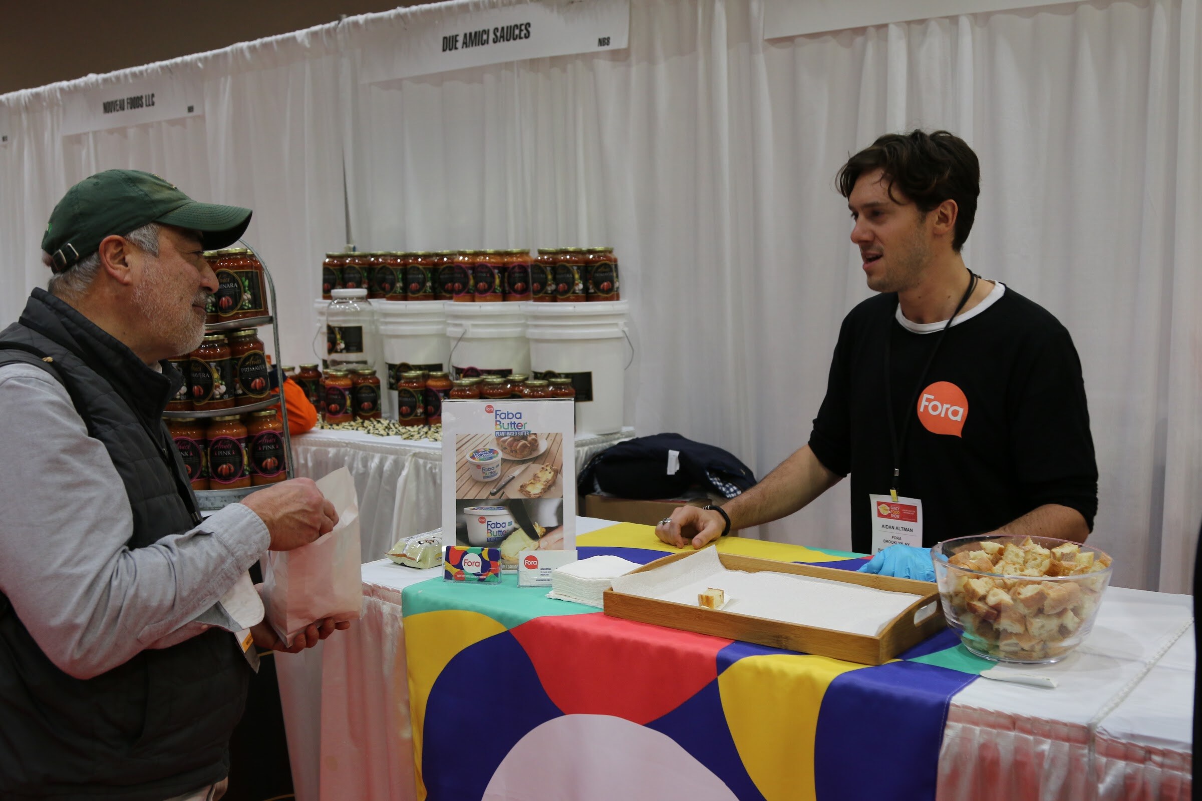 A man in a black shirt with a Fora logo at a booth counter with a visitor in a green cap.