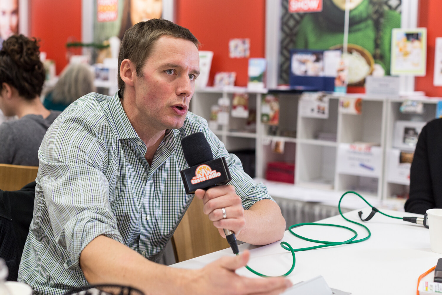 Man in a green checked shirt holding a microphone with exhibit booths behind.