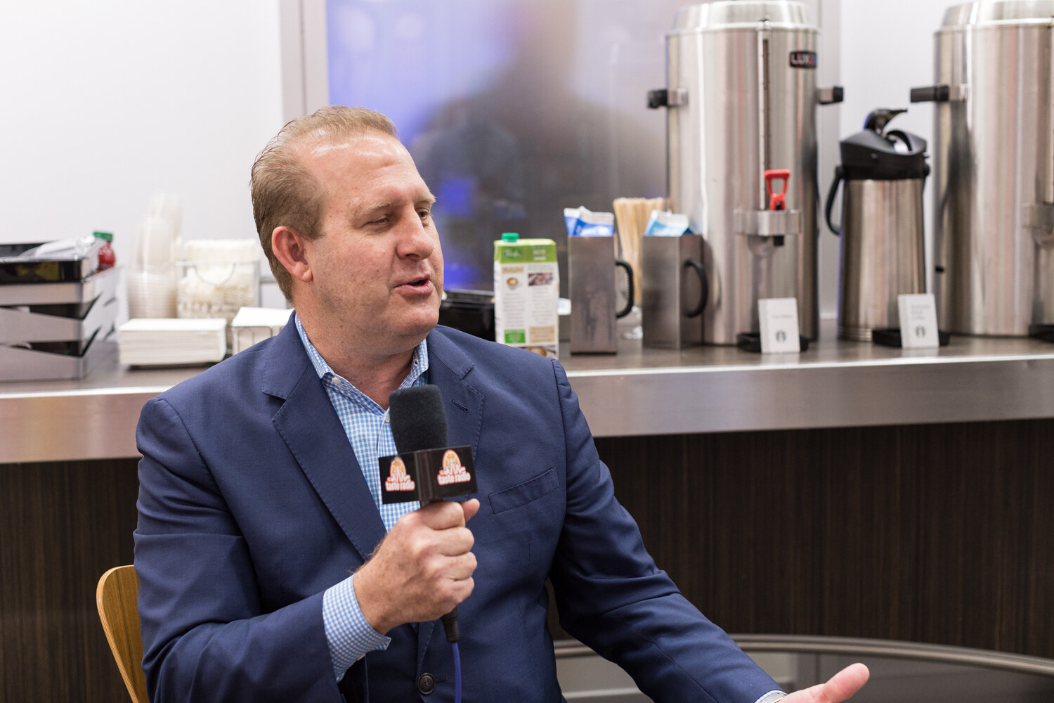 Man in a blue blazer holding a microphone in front of coffee urns.