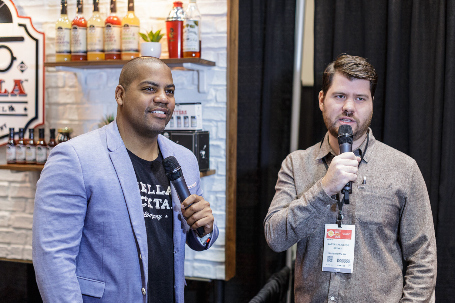 Two men holding microphones in front of a bar-style booth with a Hella Cocktail shirt visible.
