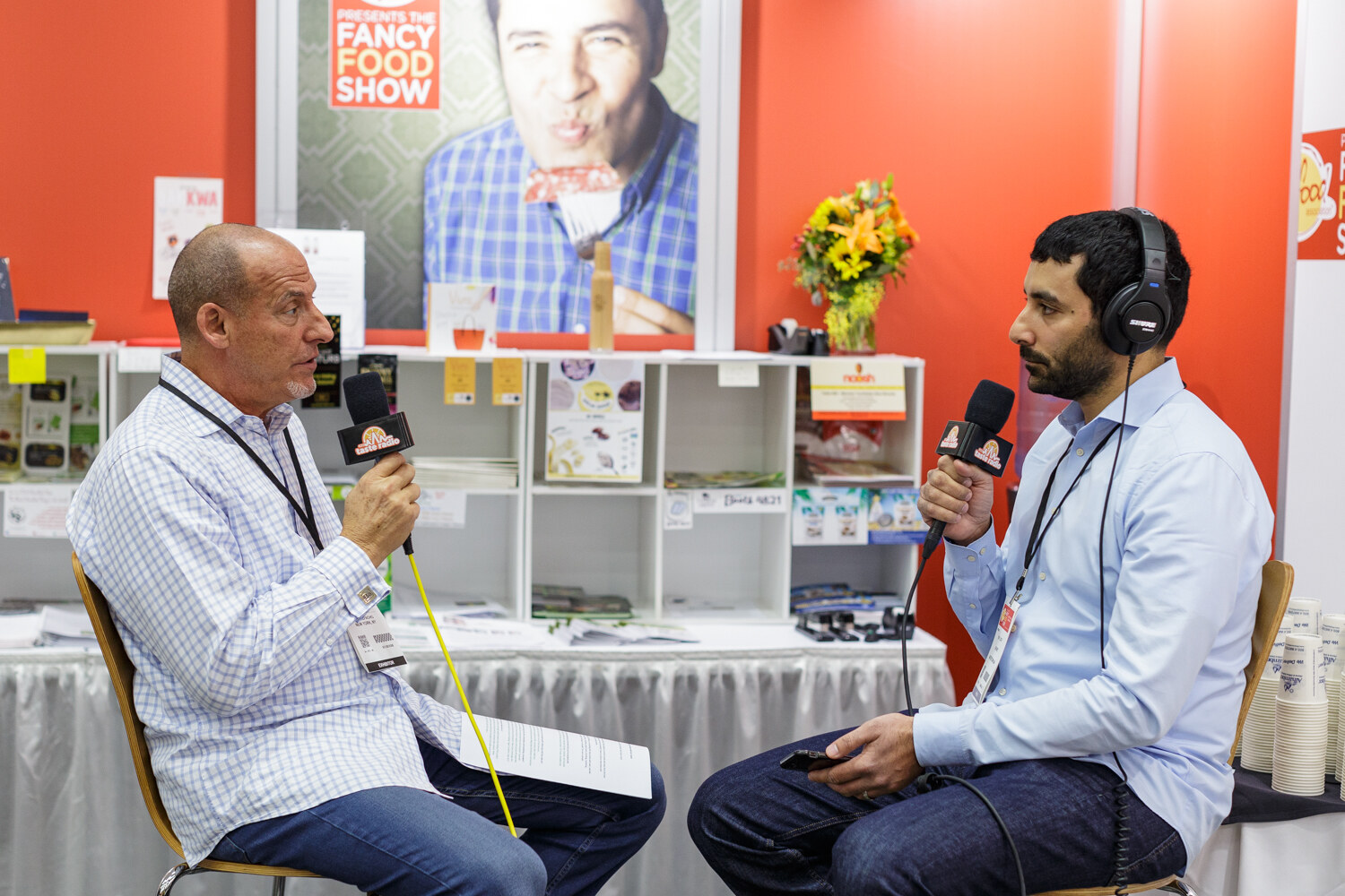 A man in a checked shirt speaks into a microphone while another man wearing headphones listens.